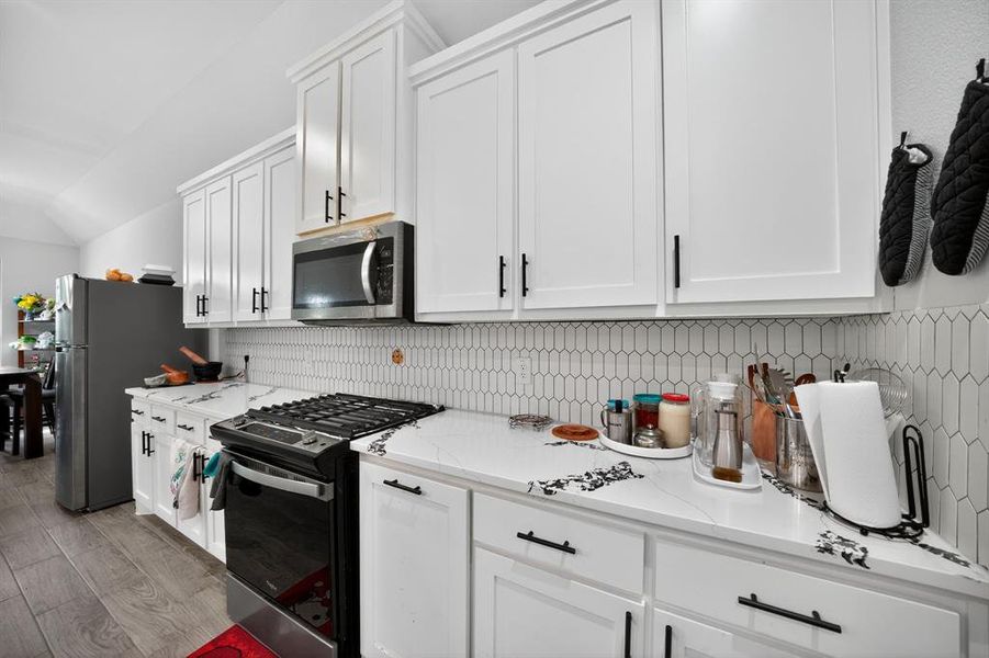 Kitchen with stainless steel appliances, white cabinetry, backsplash, light wood finished floors, and vaulted ceiling