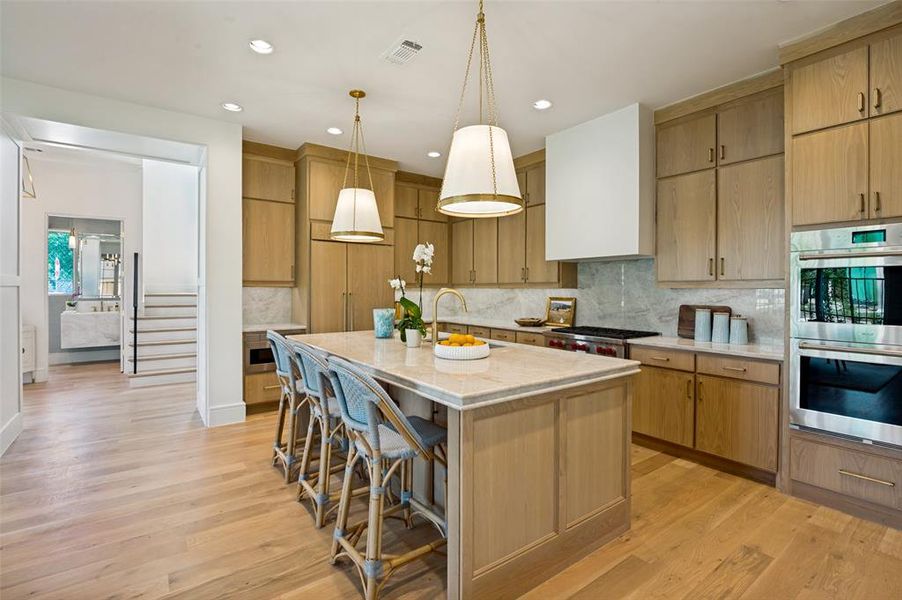 Spacious kitchen featuring light wood cabinetry, a large center island with seating, integrated stainless steel appliances, and wood-finish flooring