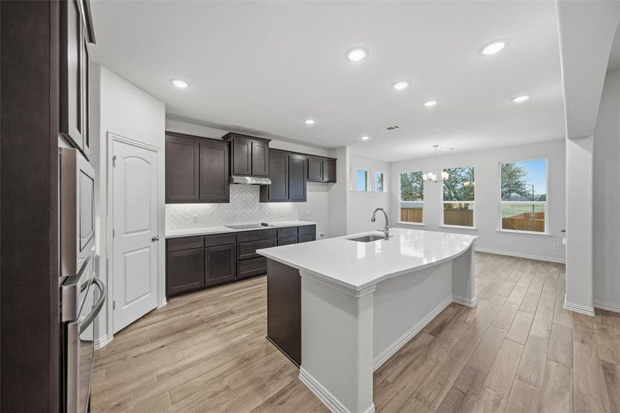 Kitchen featuring a kitchen island with sink, light wood finished floors, suspended lighting, tasteful backsplash, and light stone counters