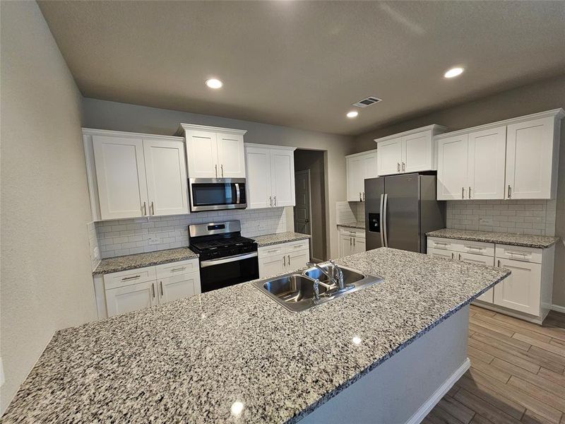 Kitchen with white cabinetry, open floor plan, stainless steel appliances, recess lights, granite counters, backsplash and wood looking tiled floors.