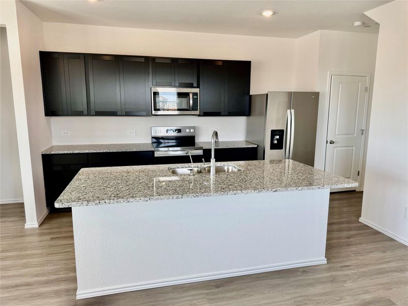 Kitchen featuring dark cabinetry, appliances with stainless steel finishes, a center island with sink, light stone counters, and light wood finished floors