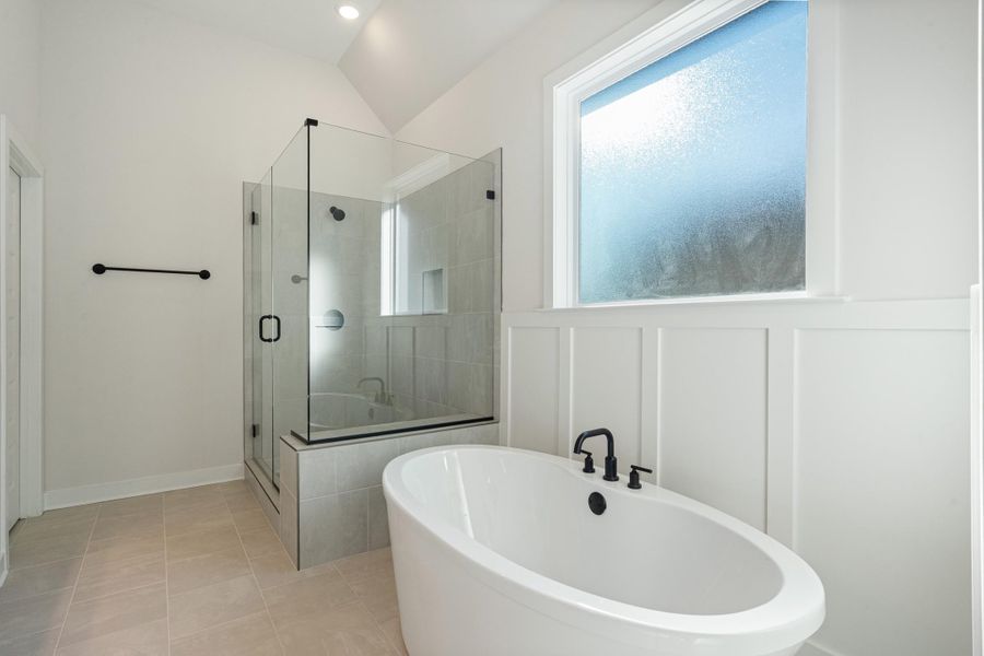Freestanding oval bathtub with matte black fixtures, glass-enclosed shower with tile surround, board and batten wall paneling, vaulted ceiling with recessed lighting, and tile flooring