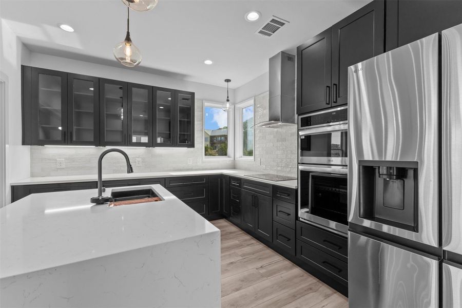 Kitchen featuring glass fronted cabinets, dark cabinets, stainless steel appliances, light stone countertops, and backsplash
