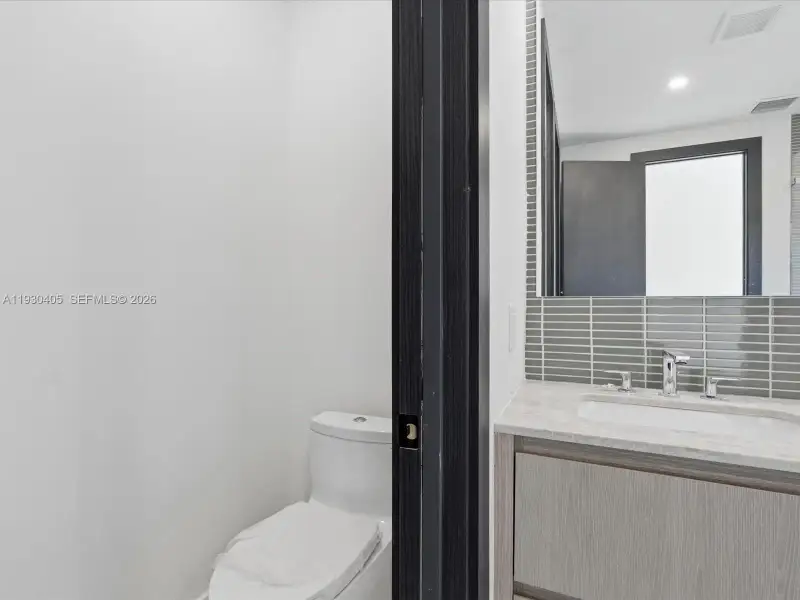 Bathroom featuring a wood-finish vanity, stone-composite countertop with an integrated basin, chrome widespread faucet, and a horizontal-tile backsplash extending to a mirror Bathroom featuring a wood-finish vanity, stone-composite countertop with an integrated basin, chrome widespread faucet, and a horizontal-tile backsplash extending to a mirror