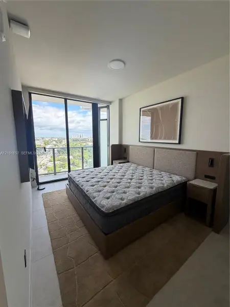 Floor-to-ceiling glass sliding doors opening to a balcony, tile flooring, and a built-in wood-finish headboard with integrated side tables