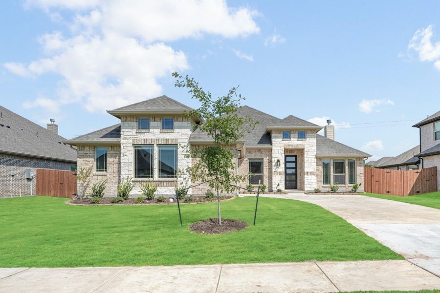 Two-story home with stone and brick exterior, large windows, and manicured front lawn with young tree