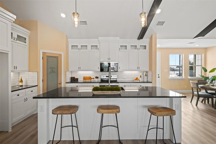 Modern kitchen featuring a large central island with seating, white shaker cabinetry, black countertops, and a subway tile backsplash Modern kitchen featuring a large central island with seating, white shaker cabinetry, black countertops, and a subway tile backsplash
