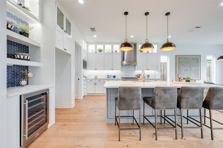 Kitchen featuring tasteful backsplash, beverage cooler, an island with sink, glass insert cabinets, and a kitchen bar Kitchen featuring tasteful backsplash, beverage cooler, an island with sink, glass insert cabinets, and a kitchen bar