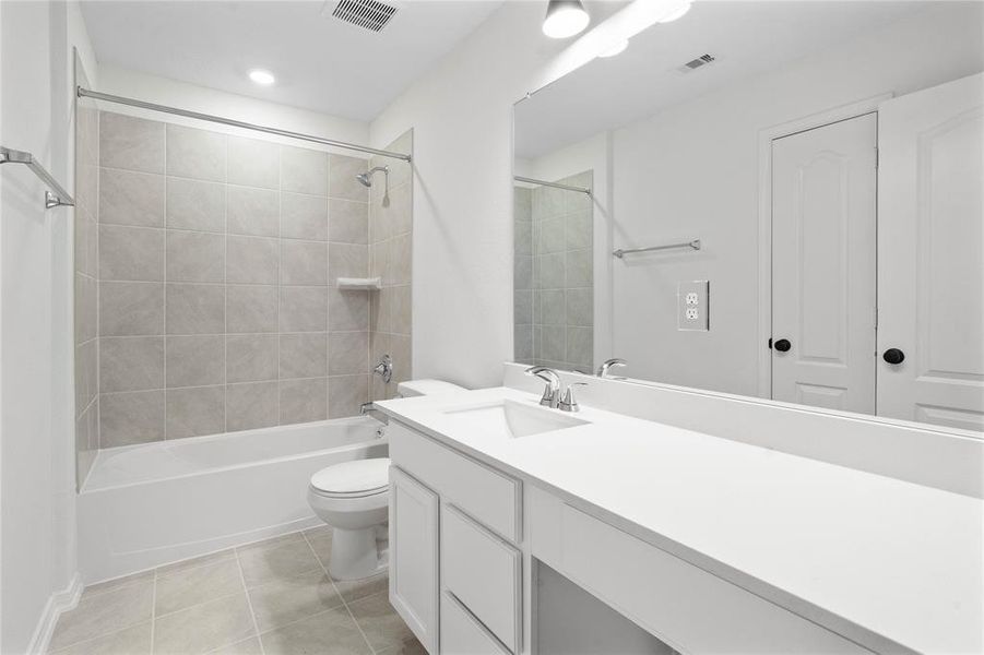 Secondary bath features tile flooring, bath/shower combo with tile surround, white stained wood cabinets, beautiful light countertops, mirror, dark, sleek fixtures and modern finishes.