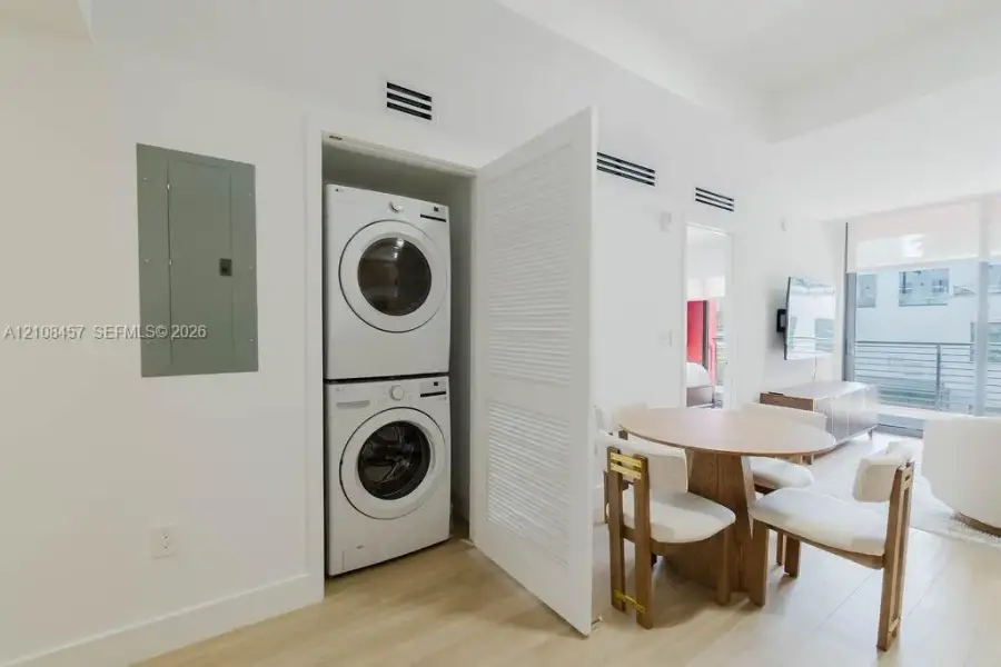 Stacked laundry center featuring a high-efficiency front-load washer and dryer enclosed by a louvered door, wood-finish flooring, and minimalist white interior walls Stacked laundry center featuring a high-efficiency front-load washer and dryer enclosed by a louvered door, wood-finish flooring, and minimalist white interior walls