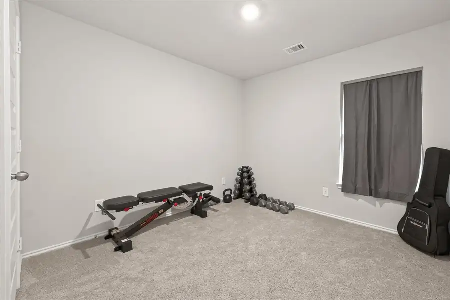 This room serves as a small home gym, featuring a weight bench, dumbbells, and carpeted flooring. A window with a curtain provides natural light, making it a versatile space for exercise or other uses.