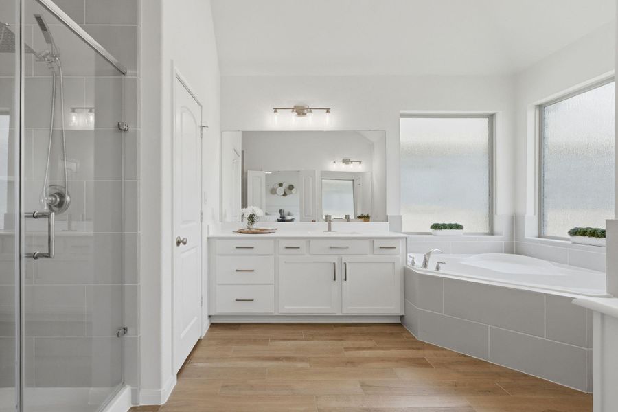 This bathroom features a modern design with a glass-enclosed shower, a large soaking tub, dual sinks with ample counter space, and sleek tile flooring. Generous natural light pours in through large frosted windows, creating a bright and inviting atmosphere.