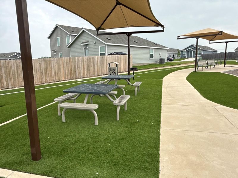 Community recreation area featuring artificial turf, a concrete pathway, shade structures, and picnic tables