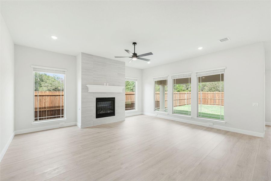 Open living room with tile-surround fireplace, ceiling fan, light wood floors, and large windows overlooking backyard
