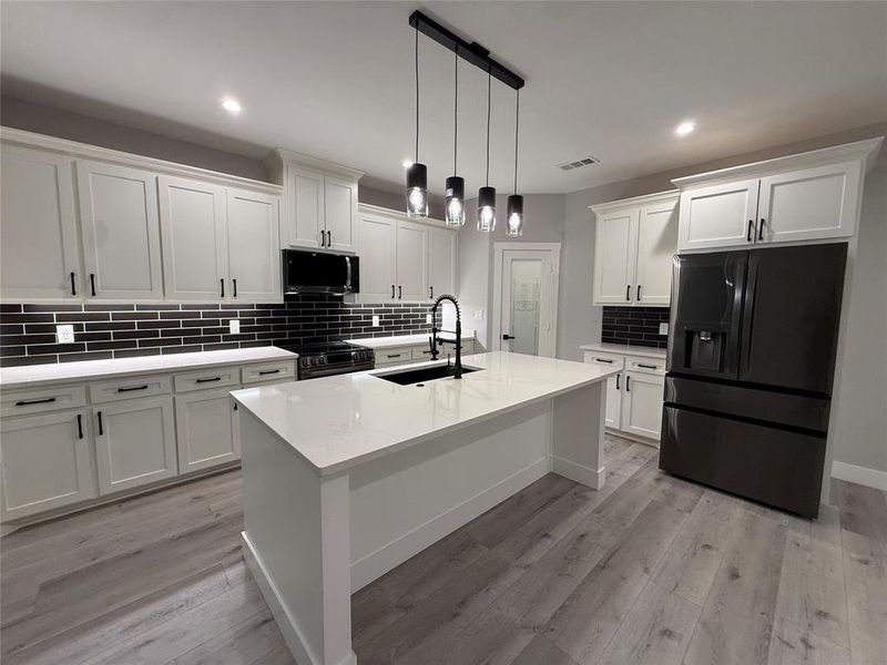 Kitchen with black appliances, light stone counters, decorative light fixtures, white cabinetry, and a center island with sink