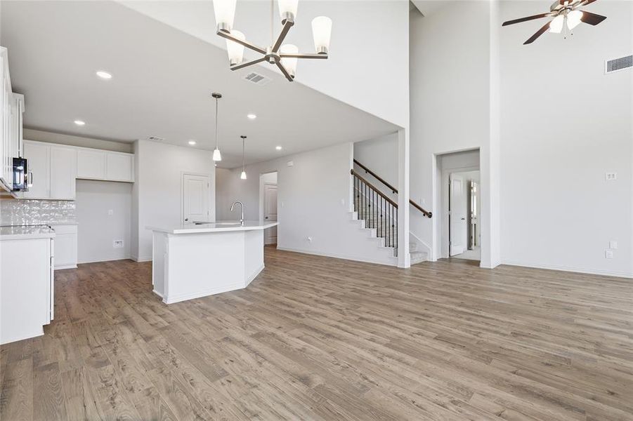 Open-concept living area featuring high ceilings, wood-finish flooring, a kitchen island with pendant lighting, white cabinetry, and a staircase with wrought iron balusters