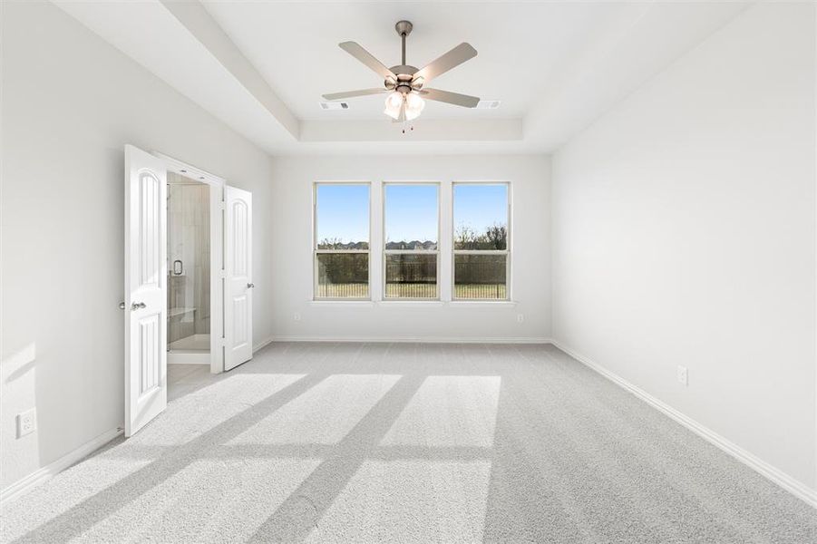 Unfurnished bedroom featuring a tray ceiling, a ceiling fan, ensuite bath, and light carpet