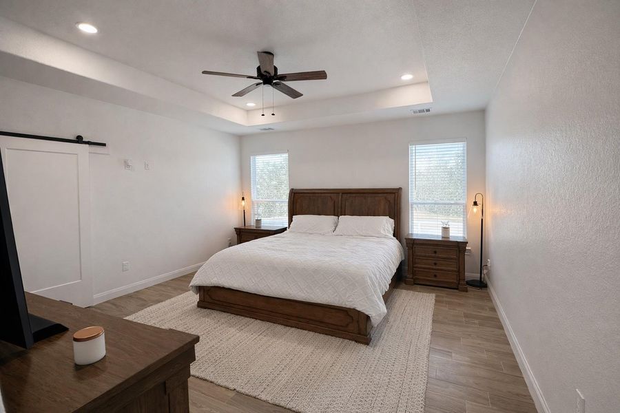 Bedroom with a barn door, wood finished floors, ceiling fan, a tray ceiling, and recessed lighting Bedroom with a barn door, wood finished floors, ceiling fan, a tray ceiling, and recessed lighting