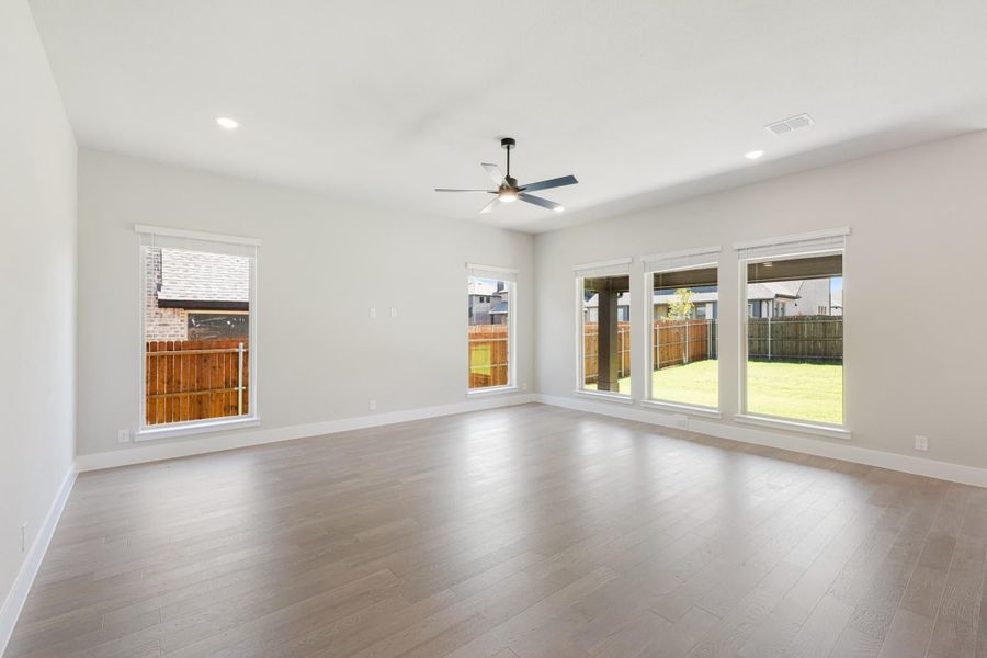 Empty living room with hardwood floors, ceiling fan, and large windows overlooking a fenced backyard