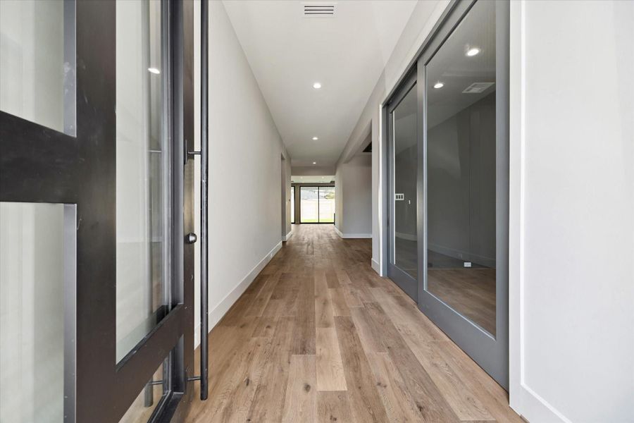 The entrance of the home makes a striking first impression with its modern glass and metal front door opening to a wide, light-filled hallway. Warm oak flooring extends the length of the space, drawing the eye toward the expansive living areas and backyard beyond, while sleek glass doors along the hall create a sense of openness and contemporary elegance.