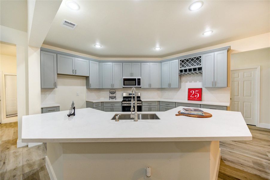 This kitchen features a modern design with light gray cabinets, a spacious white countertop island with a sink, and stainless steel appliances. The room is well-lit with recessed lighting, and there is ample storage, including a wine rack. The flooring is a warm-toned wood finish.