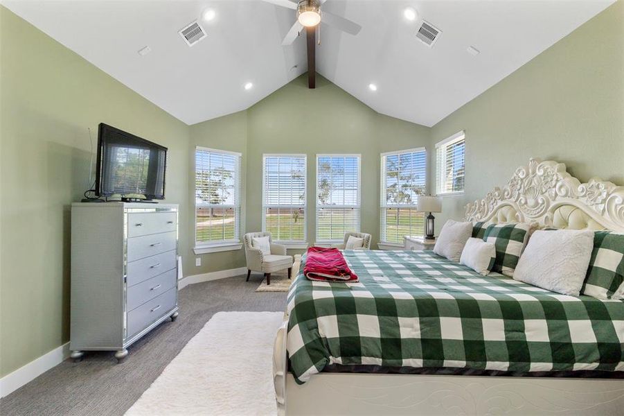 Spacious bedroom with high vaulted ceilings and large windows, allowing ample natural light. Features a stylish bed with a patterned quilt, elegant dresser, and cozy seating area.