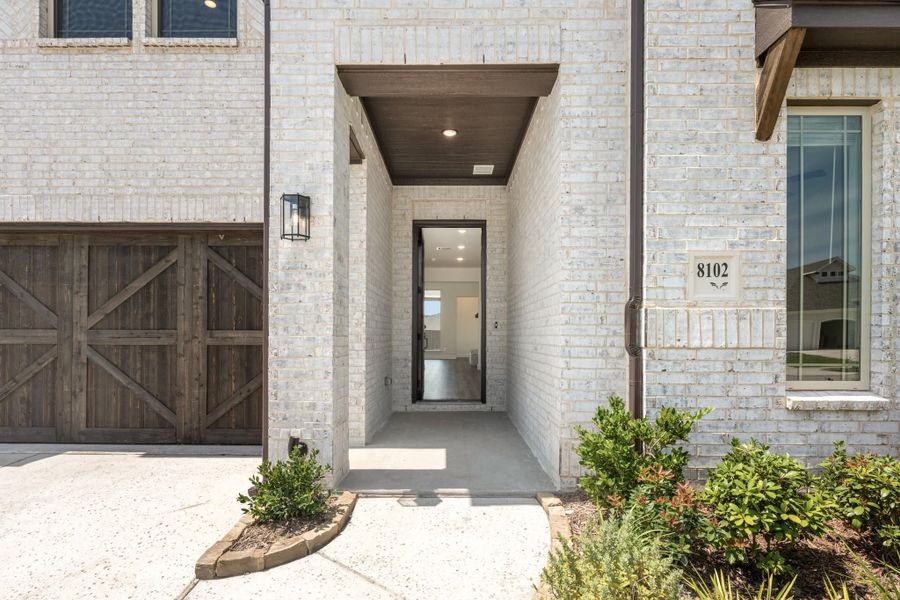 Front entry of white brick home with covered walkway, dark wood garage door, and landscaped shrubs Front entry of white brick home with covered walkway, dark wood garage door, and landscaped shrubs