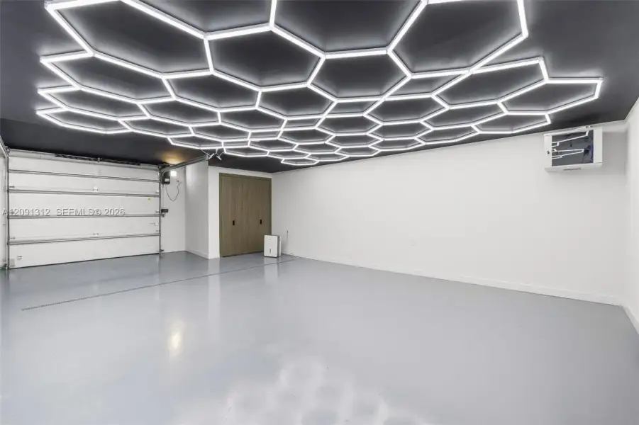 Custom ceiling with hexagonal LED light panels, smooth-finish gray flooring, white overhead garage door, and flat-panel wood-finish storage cabinetry Custom ceiling with hexagonal LED light panels, smooth-finish gray flooring, white overhead garage door, and flat-panel wood-finish storage cabinetry