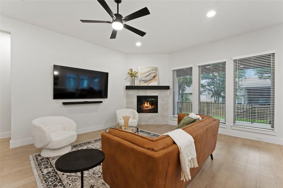 Living area featuring a corner fireplace with a tile surround and dark mantel, wood-finish flooring, a ceiling fan with integrated lighting, and multiple windows with blinds
