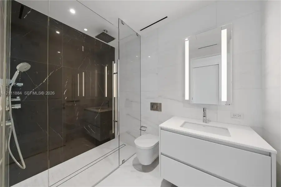 Bathroom featuring a walk-in shower with dark tile surround, a wall-mounted toilet, a floating vanity with a stone countertop, and an integrated LED mirror Bathroom featuring a walk-in shower with dark tile surround, a wall-mounted toilet, a floating vanity with a stone countertop, and an integrated LED mirror