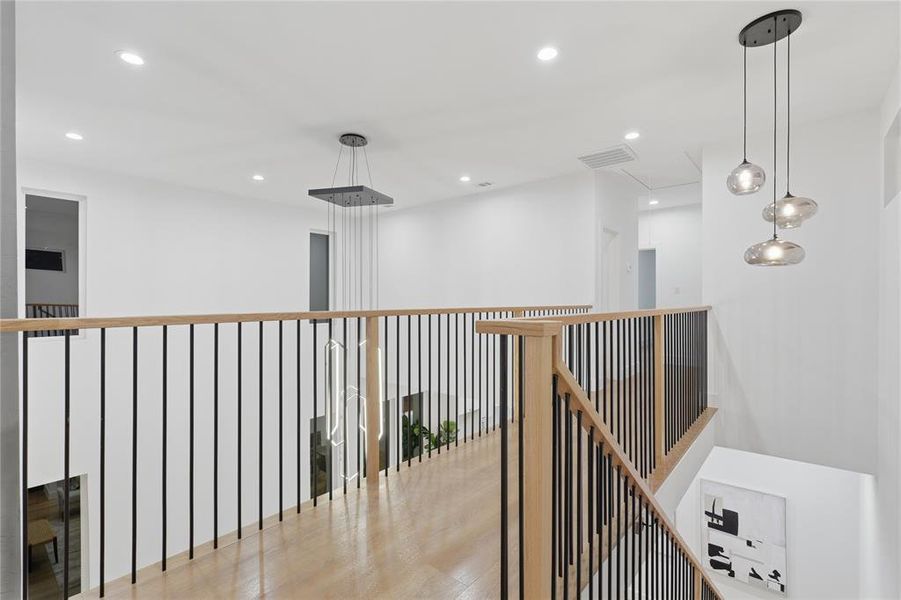 Upper-level hallway featuring wood-finish flooring, light wood handrails with black metal balusters, and recessed lighting Upper-level hallway featuring wood-finish flooring, light wood handrails with black metal balusters, and recessed lighting
