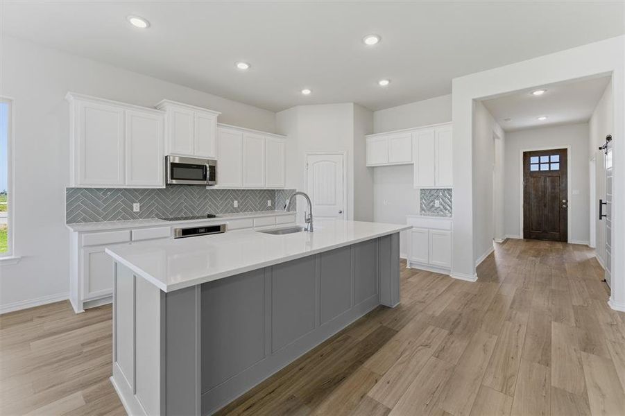 Open-concept kitchen featuring an expansive center island with a white countertop and gray paneling, stainless steel appliances, a gray herringbone tile backsplash, white shaker-style cabinetry, and wood-finish flooring