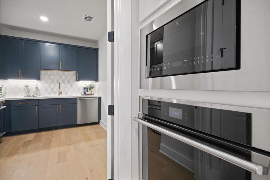 Caterers Kitchen featuring appliances with stainless steel finishes, light wood-type flooring, tasteful backsplash, blue cabinetry, and light stone countertops