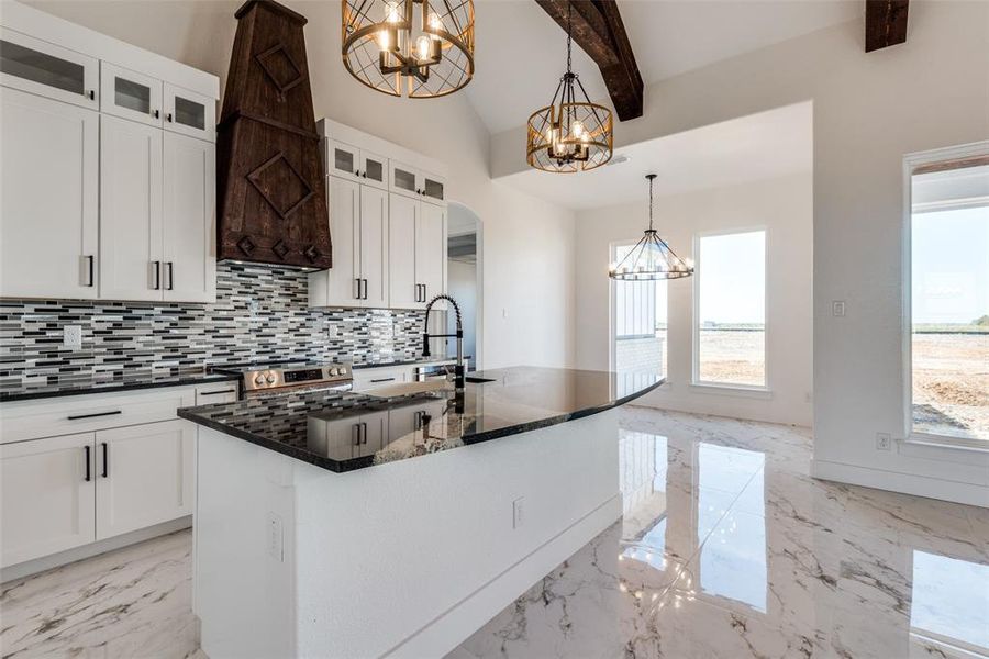 Kitchen featuring dark stone counters, a center island with sink, lofted ceiling with beams, decorative light fixtures, and white cabinets