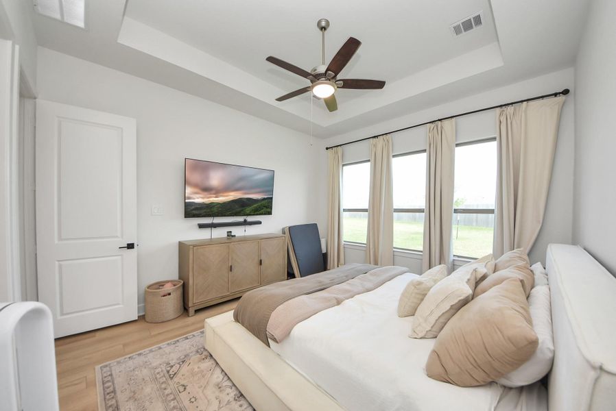 Bright and inviting bedroom featuring a tray ceiling with a ceiling fan, large windows with serene views, and a cozy bed adorned with plush pillows.