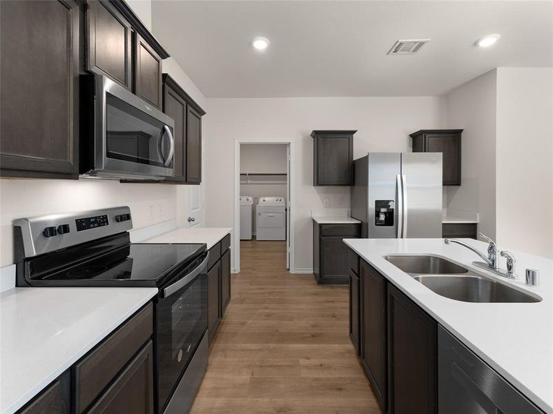 Kitchen with appliances with stainless steel finishes, sink, washing machine and clothes dryer, dark brown cabinetry, and light hardwood / wood-style floors