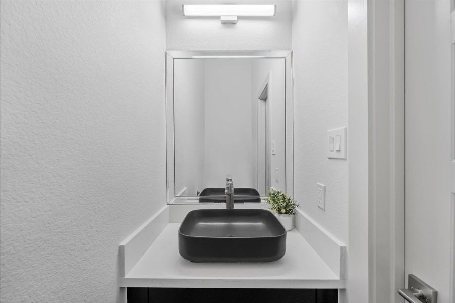 This photo showcases a modern bathroom with a sleek black vessel sink on a white countertop. A large mirror is mounted above, and the space is well-lit with a contemporary light fixture. An elegant, minimalistic design with a small plant for a touch of greenery. This photo showcases a modern bathroom with a sleek black vessel sink on a white countertop. A large mirror is mounted above, and the space is well-lit with a contemporary light fixture. An elegant, minimalistic design with a small plant for a touch of greenery.