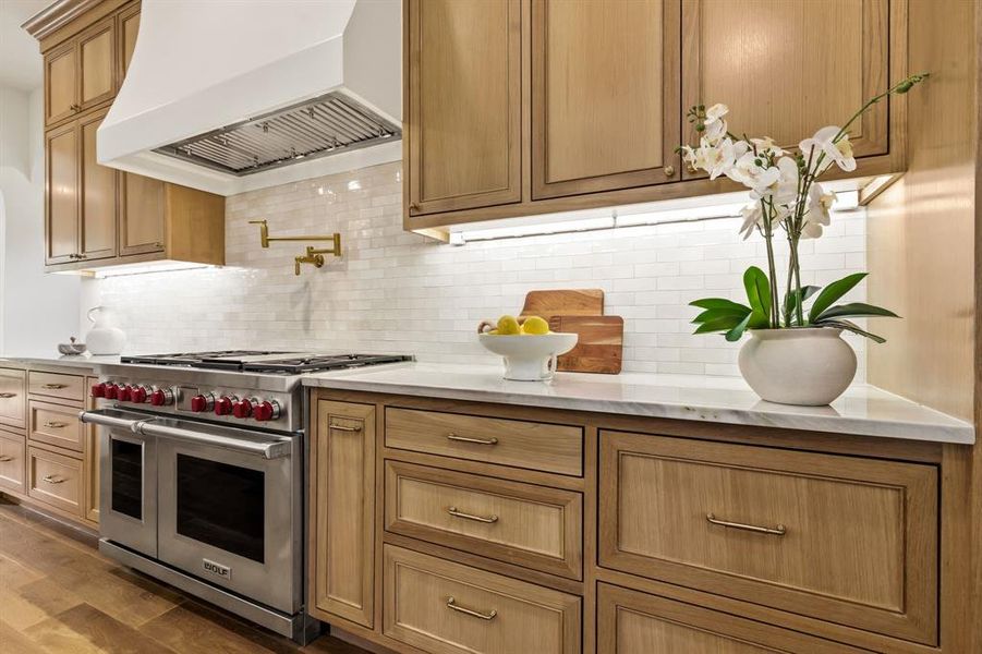 Gourmet kitchen with professional-grade stainless steel range, white subway tile backsplash, light wood cabinetry, white stone countertops, and a brass pot filler faucet