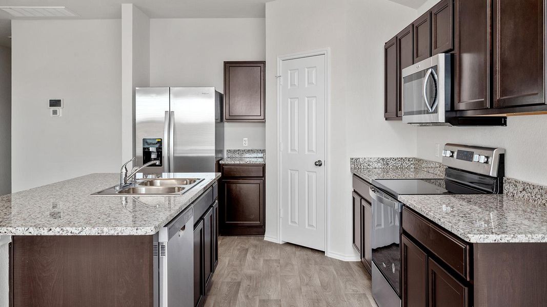 Kitchen featuring stainless steel appliances, dark wood finish cabinets, an island with sink, light wood-style floors, and light stone countertops