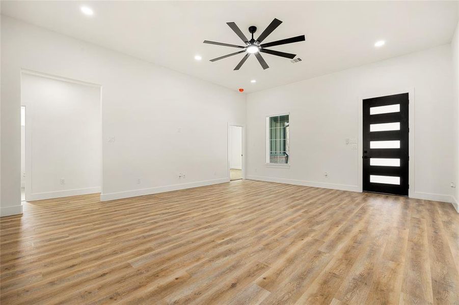 This room features modern wood flooring, a sleek black ceiling fan, and ample lighting. The space is bright with white walls, a contemporary black front door, and a window for natural light. Ideal for a modern, open-concept living area.