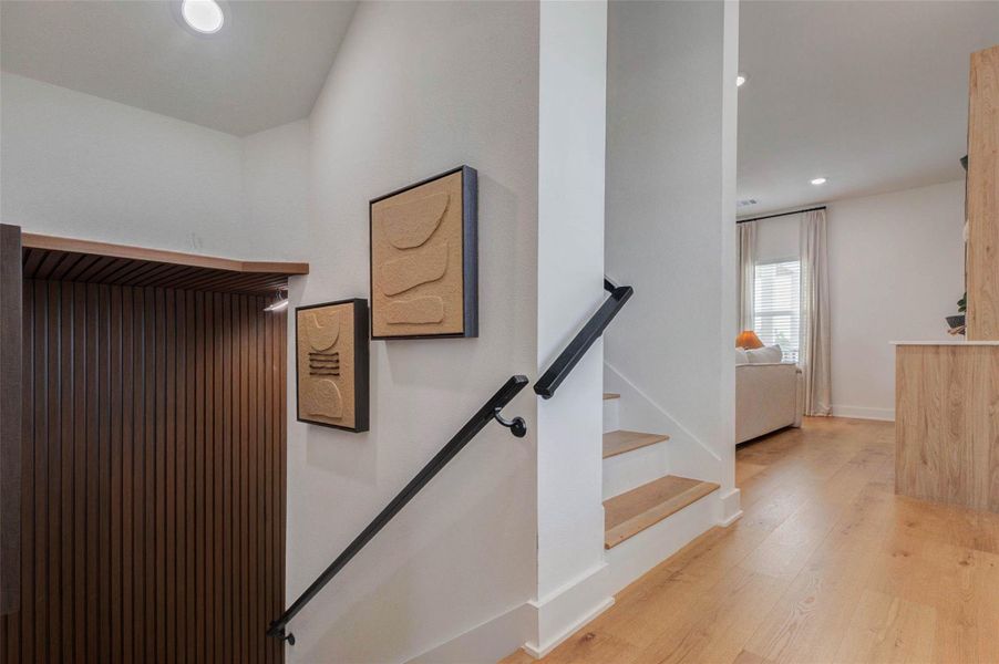 A thoughtfully designed stairway introduces modern architectural detail with warm wood treads, crisp white walls, and sleek black handrails. Textured artwork adds visual interest along the stair wall, while vertical wood slats create a distinctive feature at the lower level. The open hallway connects smoothly to the adjacent living space, and recessed lighting keeps the area bright, clean, and welcoming.