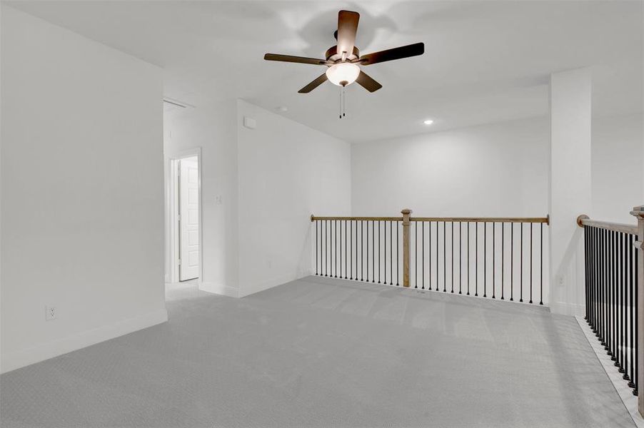 Spacious landing area featuring light grey carpet, a ceiling fan, and a wooden railing with black balusters