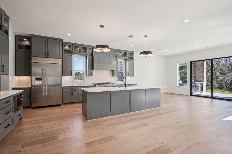 This spacious kitchen features modern gray cabinetry, a large island with a sink, stainless steel appliances, and elegant pendant lighting. The open floor plan flows into a bright living area with large sliding doors leading to an outdoor space, perfect for entertaining.
