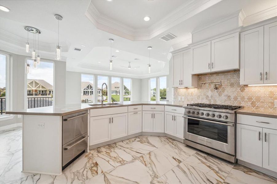 This is a spacious, modern kitchen with white cabinetry, high-end stainless steel appliances, and a stylish marble floor. It features a large island, elegant pendant lighting, and a view of the water beyond large windows.