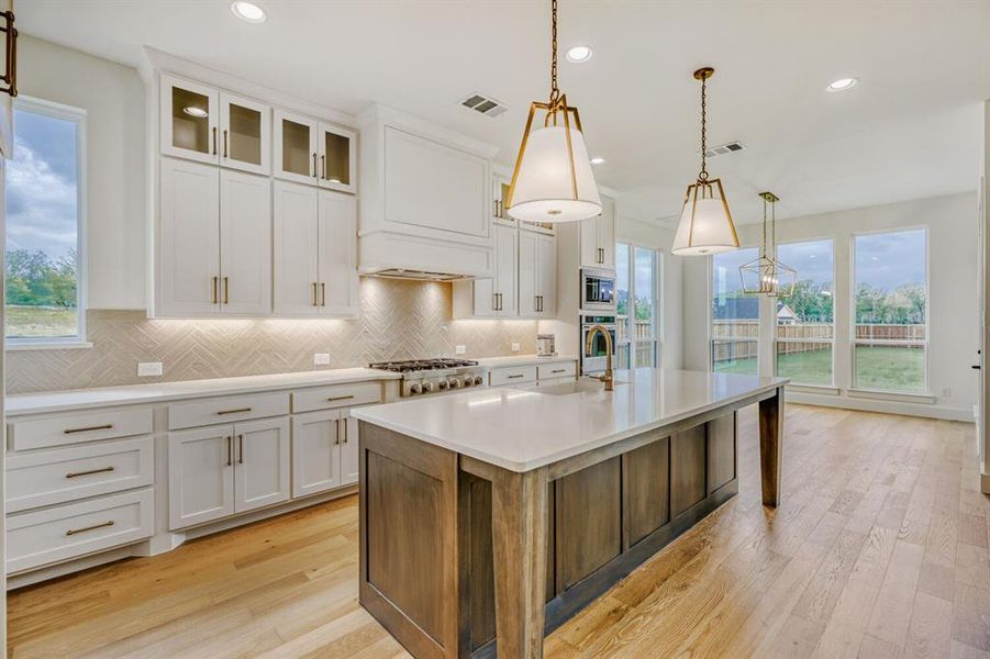 Kitchen with two tone cabinetry, hanging light fixtures, an island with sink, light wood-type flooring, and glass insert cabinets