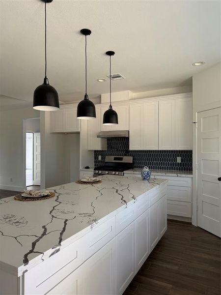 This is a modern kitchen featuring white cabinetry, a marble countertop with a striking vein pattern, stylish black pendant lights, and a bold hexagonal tile backsplash. The room is well-lit with natural light and includes contemporary finishes.