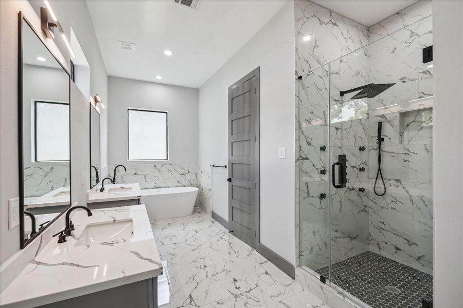 The primary bathroom is anchored by dual vanities, a glass-enclosed shower, and a soaking tub set beneath a window. Coordinated tilework ties the space together, accented by dark fixtures for contrast.
