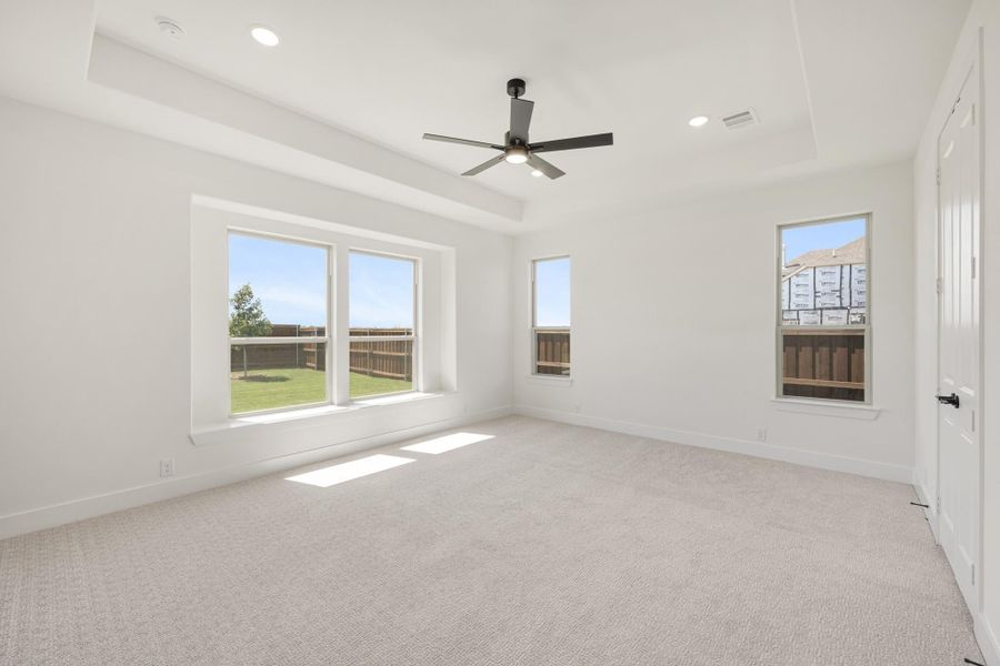Empty bedroom with tray ceiling, ceiling fan, carpet flooring, and multiple windows overlooking a fenced backyard