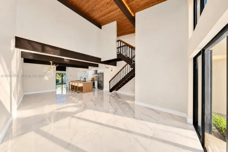 High-ceiling interior featuring a cedar wood-plank ceiling with exposed structural beams, large-format tile flooring, a geometric multi-arm light fixture, and a floating staircase with bronze metal railings High-ceiling interior featuring a cedar wood-plank ceiling with exposed structural beams, large-format tile flooring, a geometric multi-arm light fixture, and a floating staircase with bronze metal railings