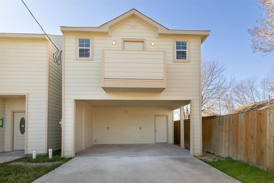 This is a two-story home with a beige exterior, featuring a covered carport. The upper level has a small balcony and two windows. The property is bordered by a wooden fence, offering some privacy.